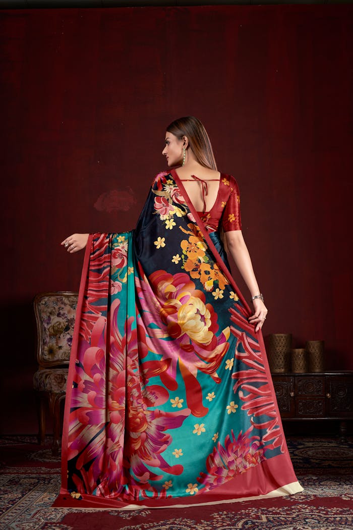 A woman in a vibrant floral saree poses indoors against a red wall, embodying traditional Indian attire.